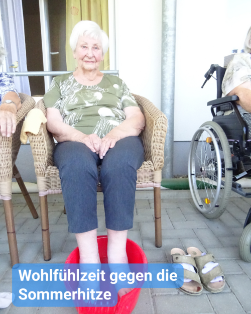 Eine ältere Frau sitzt im Korbstuhl, mit den Füßen in einer roten Wasserschüssel. Text: 'Wohlfühlzeit gegen die Sommerhitze'