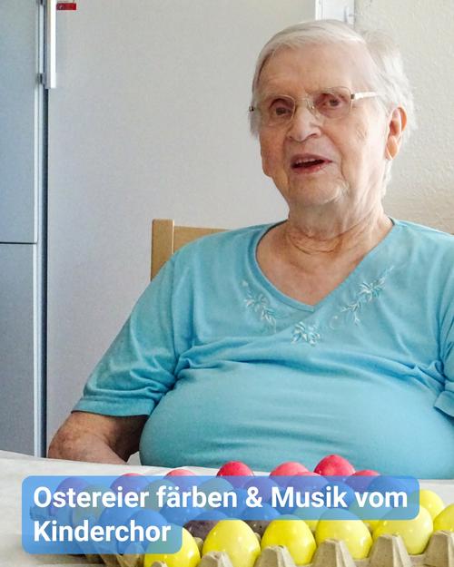 Nahaufnahme einer älteren Frau mit kurzem, hellgrauem Haar, die eine Brille trägt und ein leicht blaues, kurzärmliges Hemd anhat. Sie sitzt offenbar an einem Tisch. Ihr Ausdruck ist freundlich, und sie blickt direkt in die Kamera.  Vor ihr befinden sich zahlreiche bunt bemalte Ostereier in einem Eierkarton. Die Eier weisen verschiedene Schattierungen von Rosa, Gelb und möglicherweise weiteren Farben auf.  Ein blauer Banner am unteren Rand besagt: "Ostereier färben & Musik vom Kinderchor."